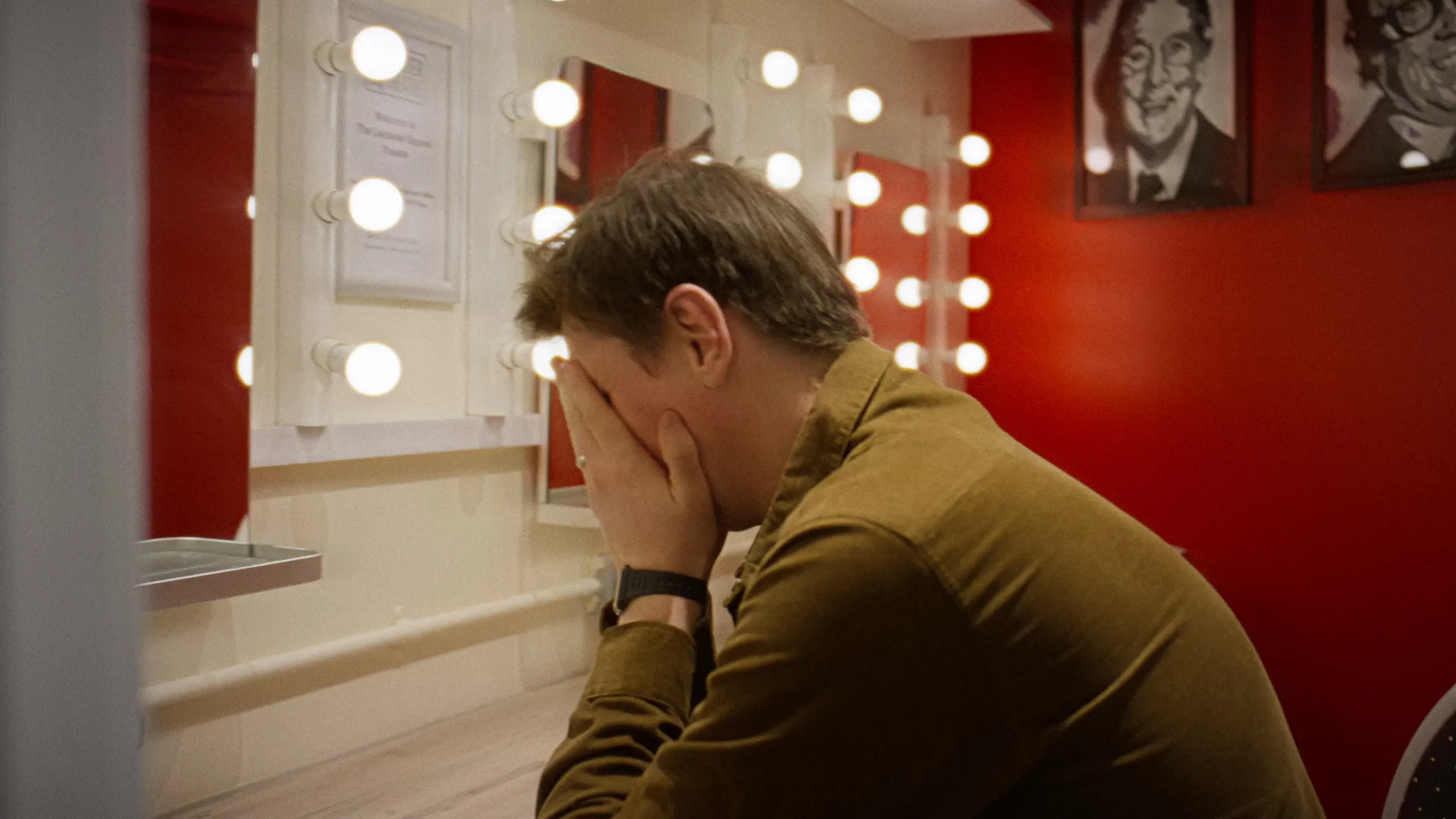 Sean McLoughlin with his face in his hands in a dressing room with a red wall and bulb mirror